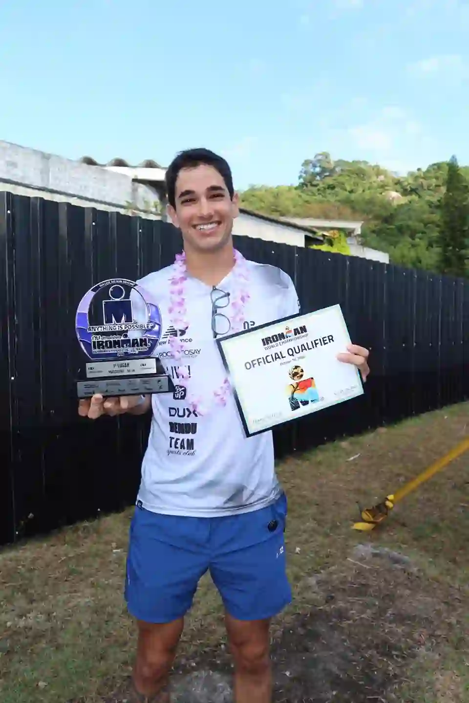Gui Vaz sorrindo, segurando o troféu de 1º lugar na categoria 30-34 e o certificado oficial de classificação para o Campeonato Mundial de Ironman em Kona, no Ironman Brasil 2026